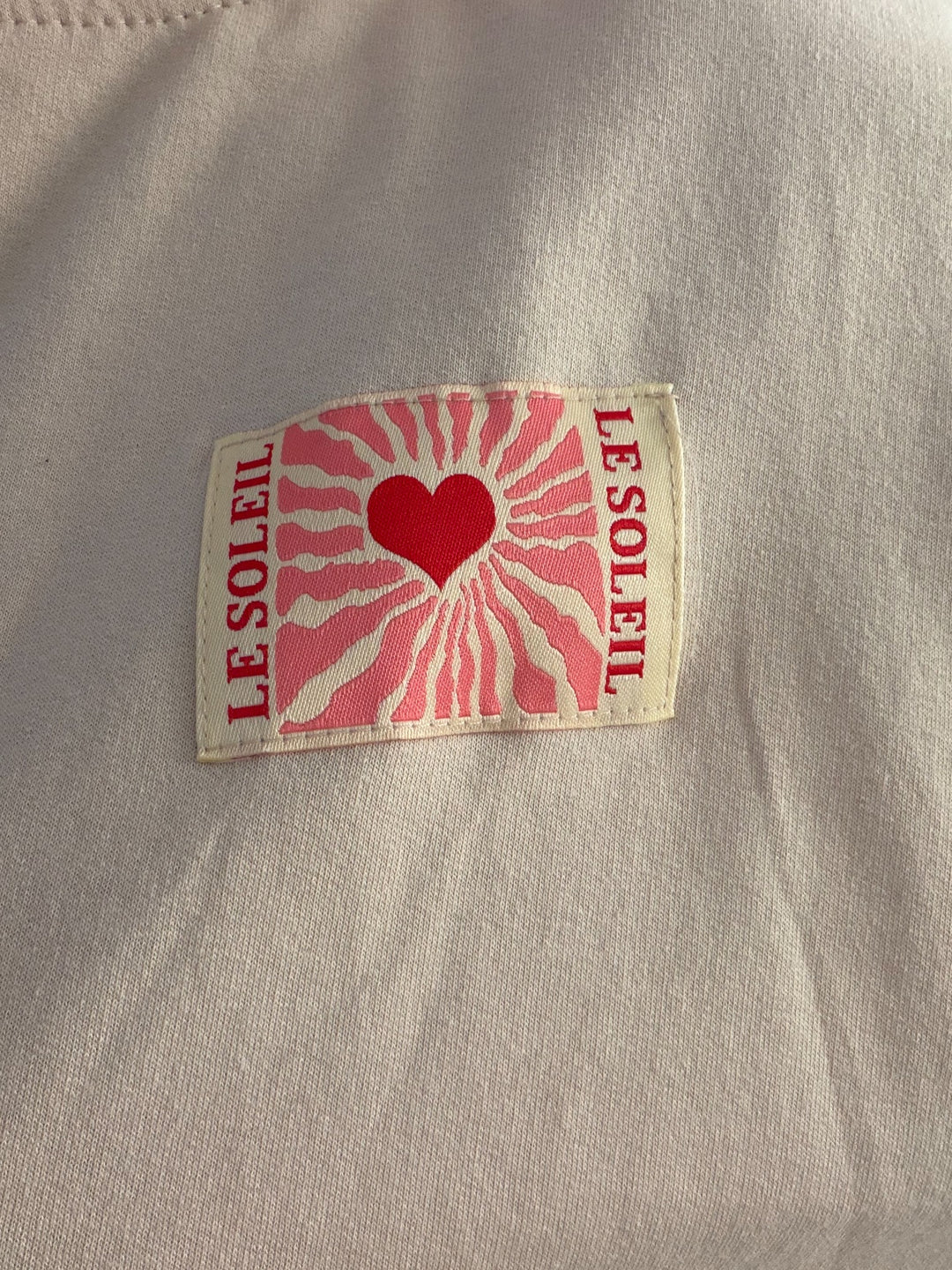 Detail des Le-Soleil-Herz-Patches auf dem beigen Langarmshirt