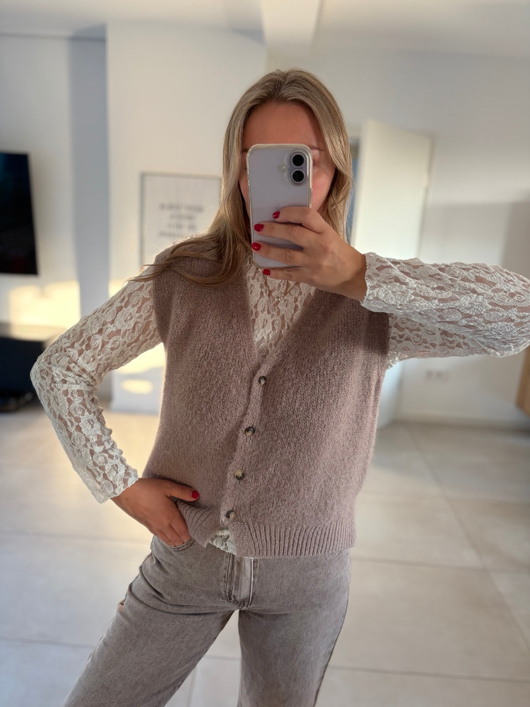 Taupefarbene Strickweste mit vier Knöpfen, kombiniert mit weißer Spitzenbluse und heller Jeans